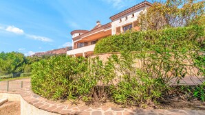 Une maison à deux étages avec un balcon, entourée d’une végétation luxuriante et sous un ciel dégagé.
