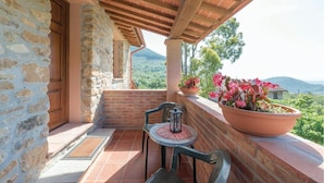 A balcony with a stone wall, a wooden door, a brick floor, two chairs, a table with a lantern, and a large potted plant with pink flowers.