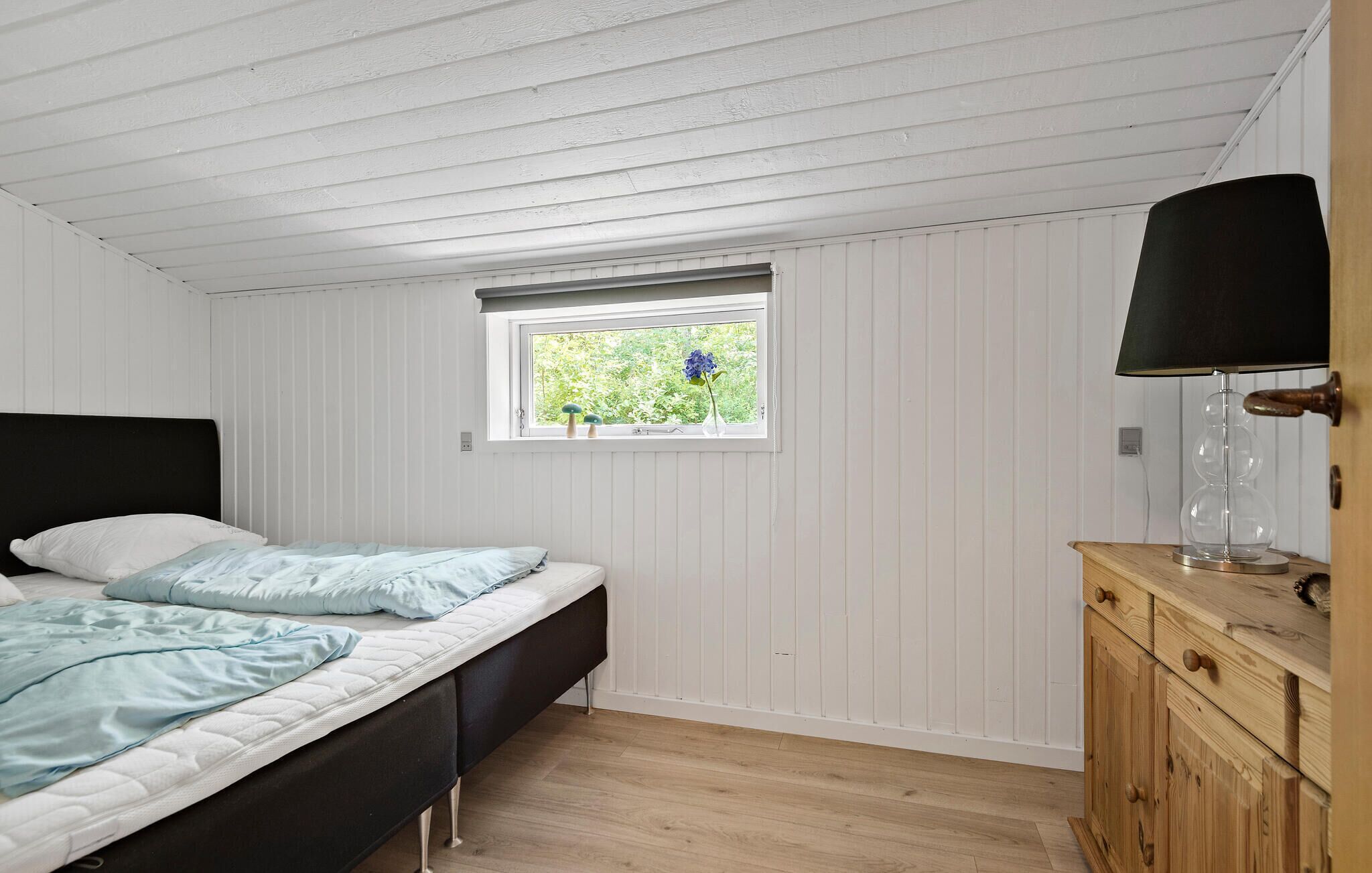 Ein Schlafzimmer mit einem Bett, einem Fenster mit Blick auf Grünflächen, einer Holzkommode und einer Lampe.