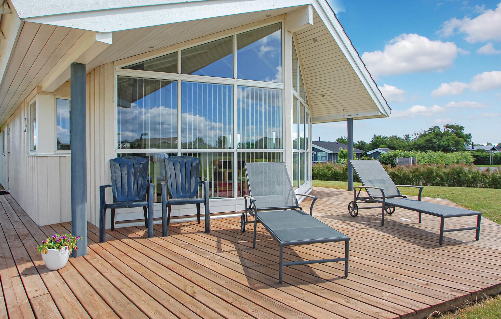 A covered outdoor deck with two lounge chairs and a table, large windows, and a white building with a sloped roof.