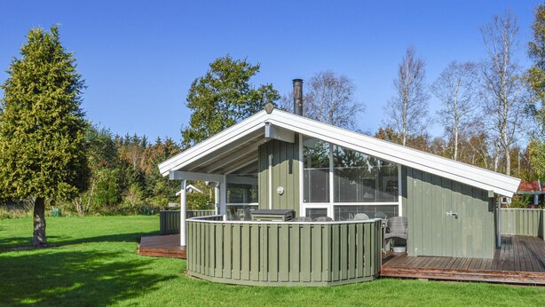 Um edifício moderno, térreo, com telhado inclinado e grandes janelas. Apresenta uma área de estar coberta ao ar livre, com um deck de madeira, uma mesa e cadeiras.