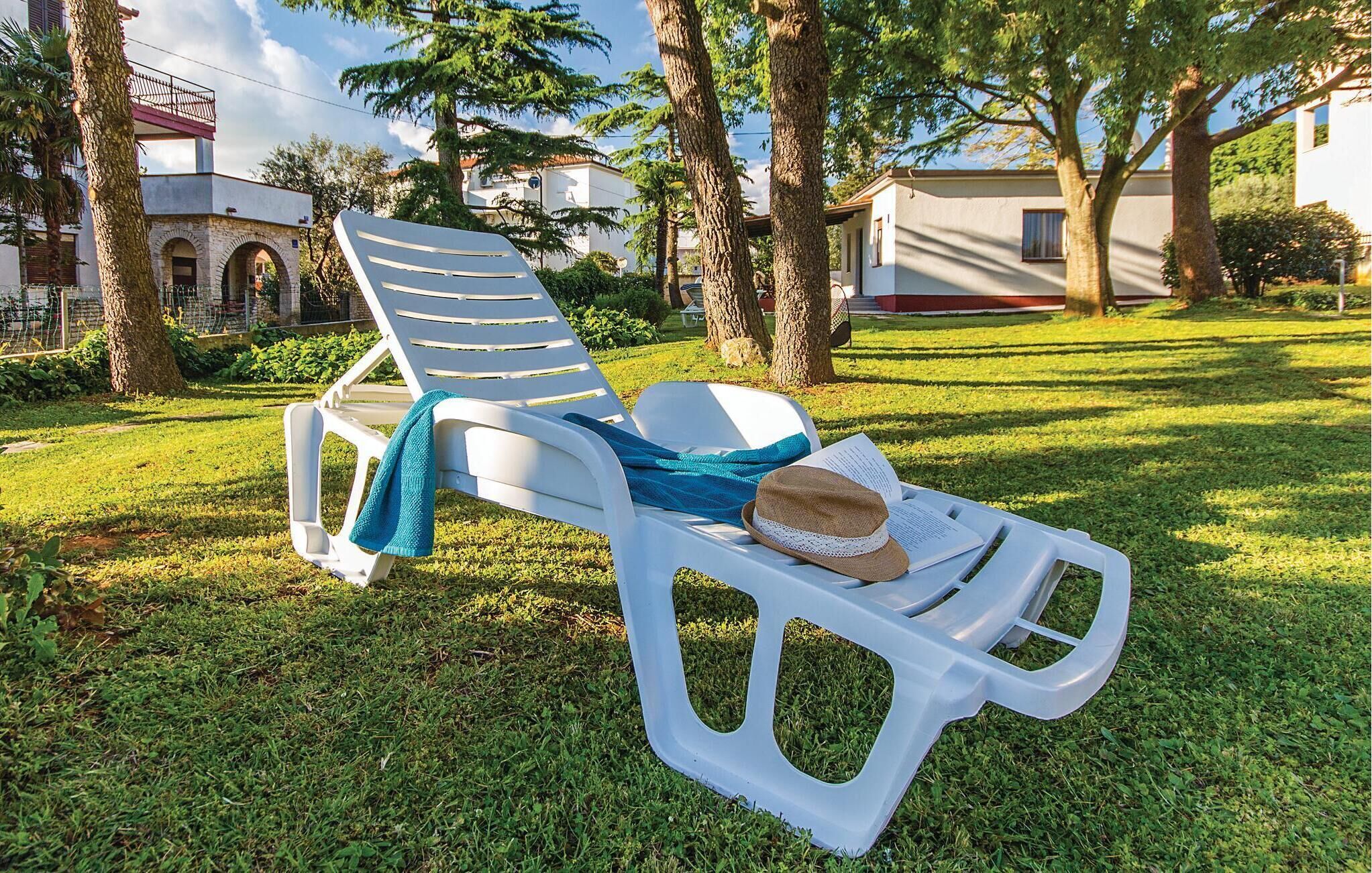A white plastic lounge chair with a towel and a straw hat on it, placed on a grassy lawn with trees and buildings in the background.