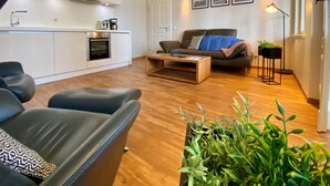Un salon moderne avec un canapé en cuir noir, une table basse en bois et un divan gris. On y trouve également un coin cuisine avec des meubles blancs et un four encastré.