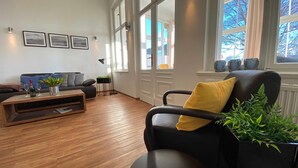 Un salon moderne avec un fauteuil en cuir noir, une table basse en bois et un canapé bleu. Des tableaux encadrés ornent le mur, ainsi qu’une lampe sur pied et une fenêtre donnant sur des arbres.