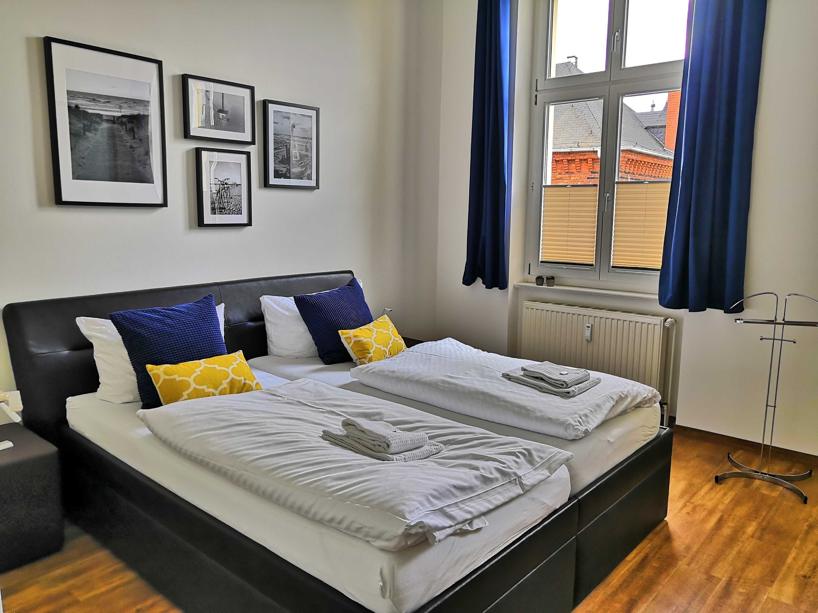 Una cama doble con sábanas blancas, almohadas azules y amarillas, un cabecero negro y una ventana con cortinas azules.