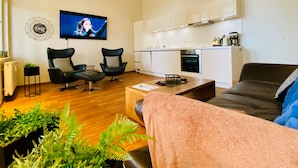 Un salon moderne comprenant un canapé, deux fauteuils, une table basse et un téléviseur. On y trouve également un coin cuisine avec des meubles blancs et une cuisinière.