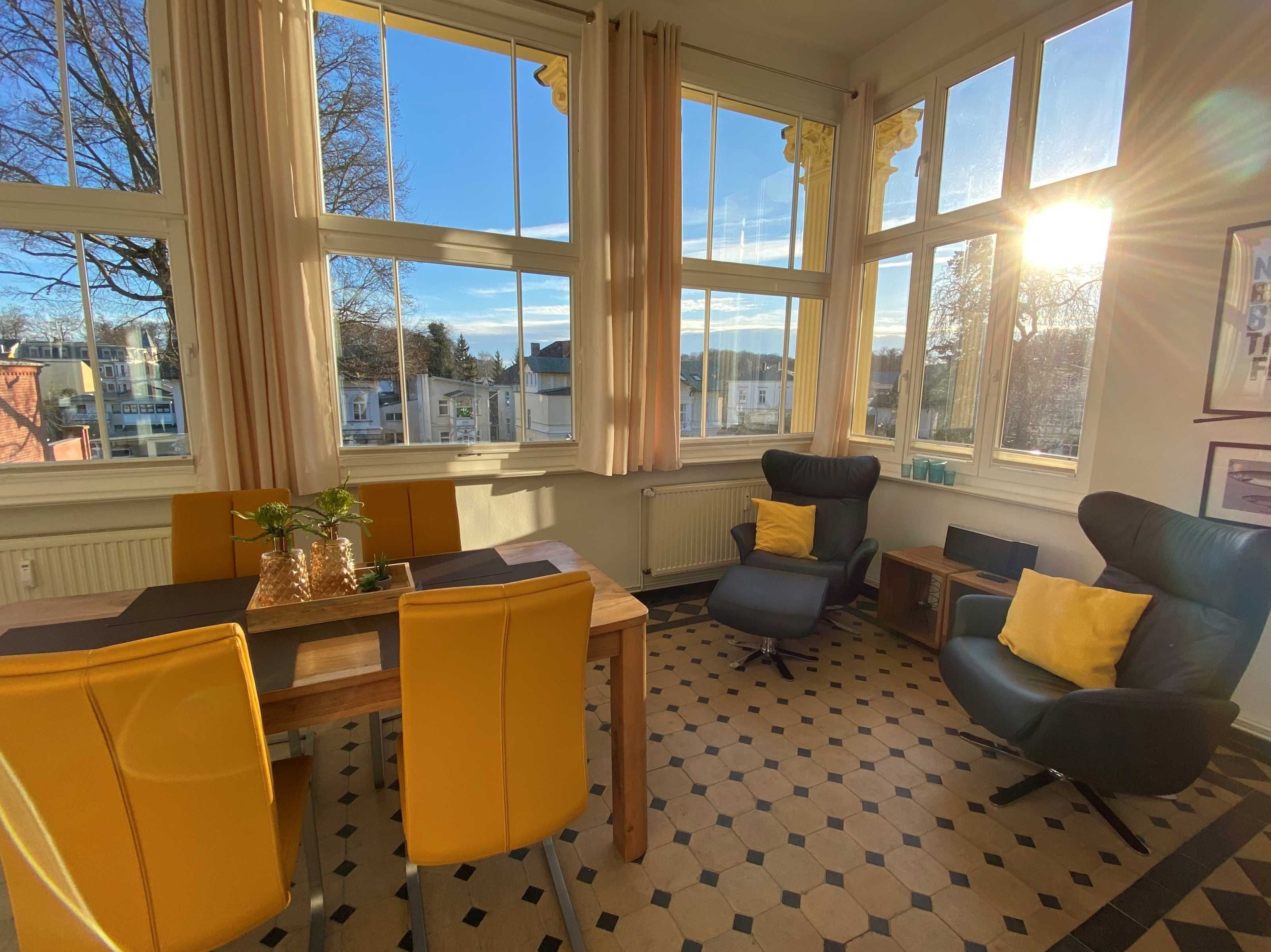 A room with a dining table, yellow chairs, a black armchair, and a blue sofa with yellow cushions. Large windows provide a view of the outside.