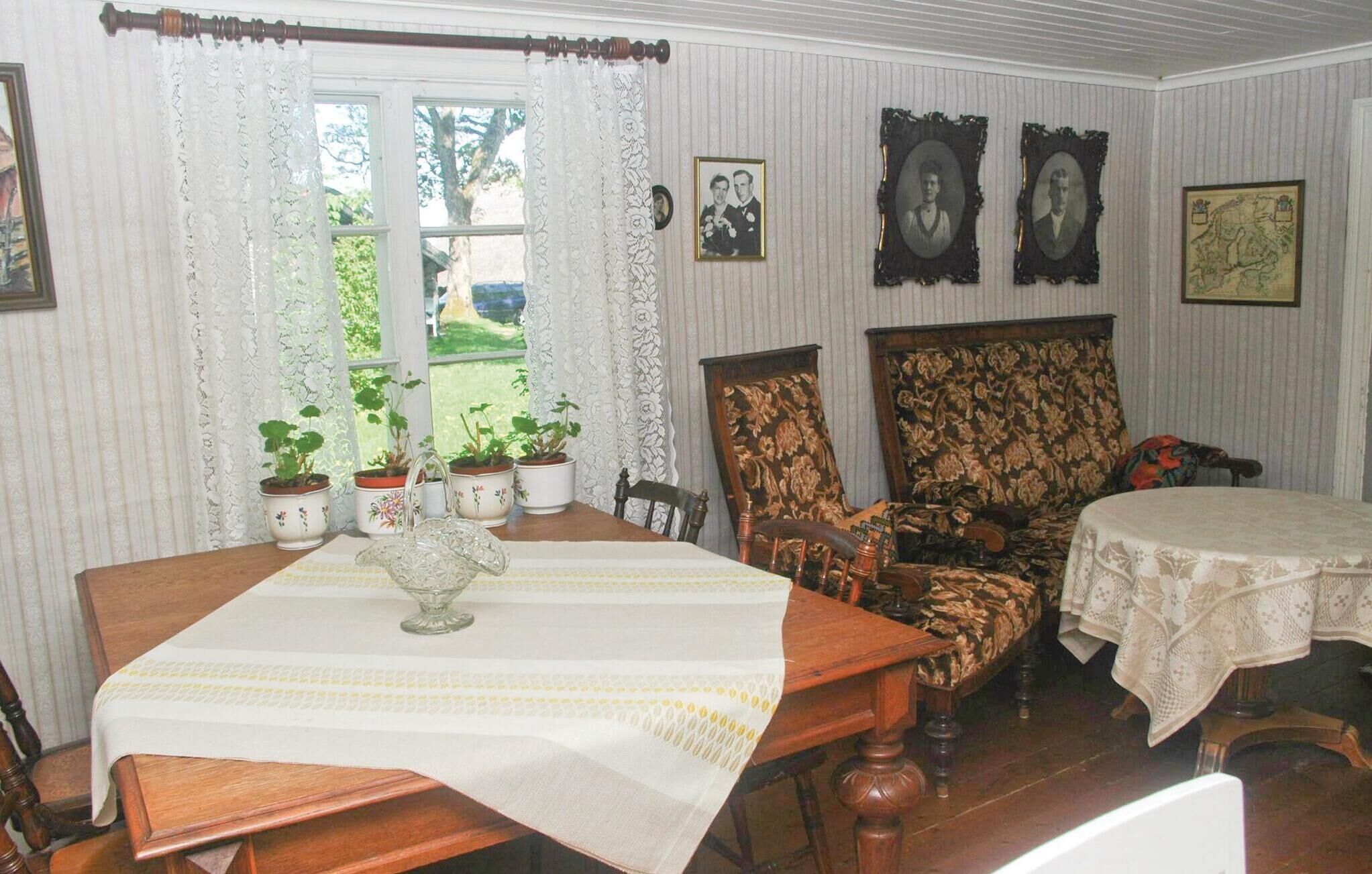 Une salle à manger avec une table en bois, des chaises et une nappe en dentelle. Des plantes en pot sont posées sur le rebord de la fenêtre et des photographies encadrées ornent le mur.