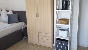 A bedroom with a bed, a wardrobe, a shelf with kitchen items, and a wall with framed pictures. - Apartment Fofftein (Friedrichskoog)