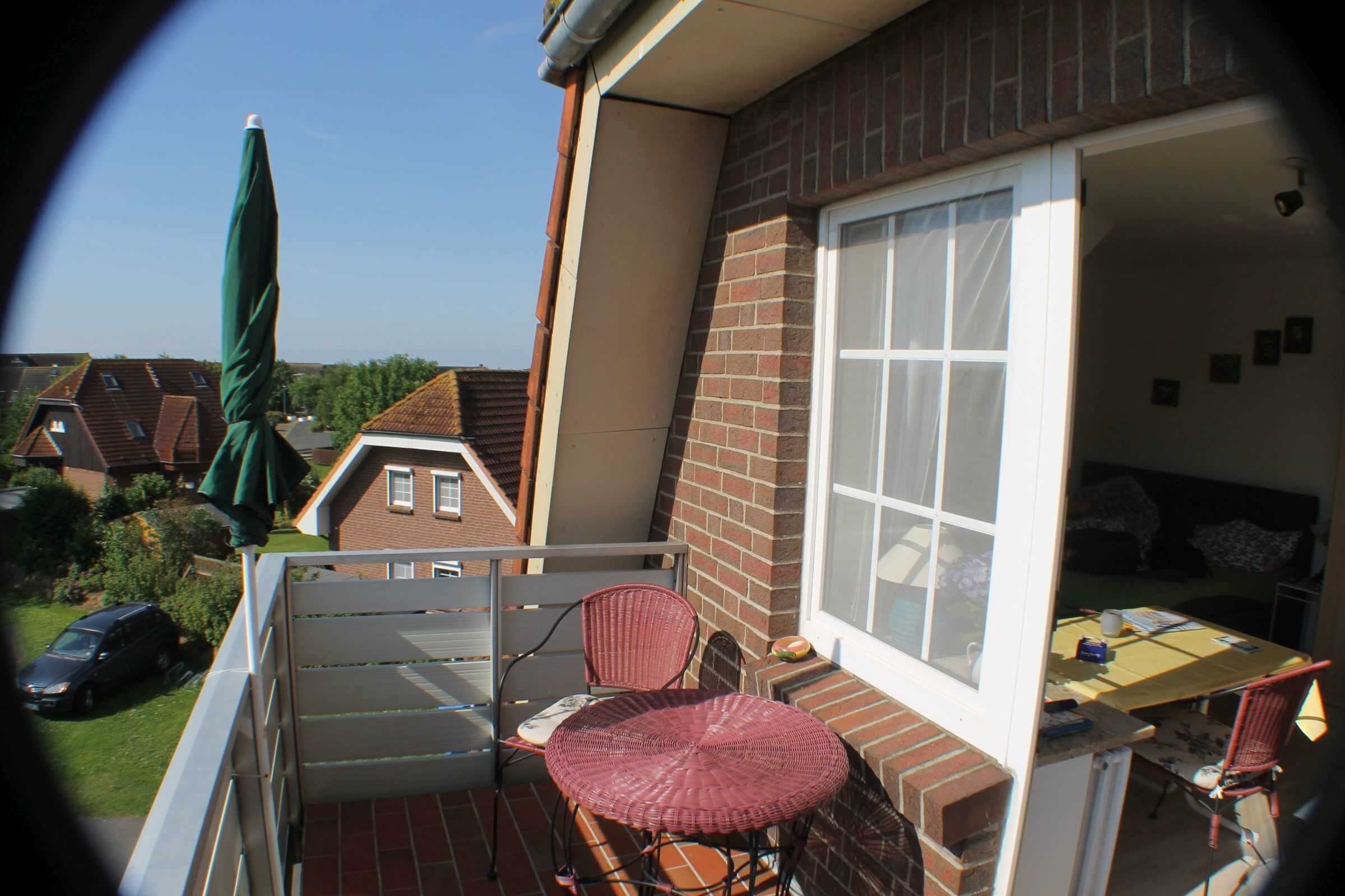 A small balcony with a wicker chair, a table, and a window overlooking a residential area.