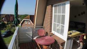 A small balcony with a wicker chair, a table, and a window overlooking a residential area. - Apartment Fofftein (Friedrichskoog)