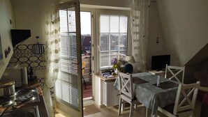 A cozy, well-lit room with a kitchen area, a dining table with chairs, a TV, and a view of the outside through large windows. - Apartment Fofftein (Friedrichskoog)