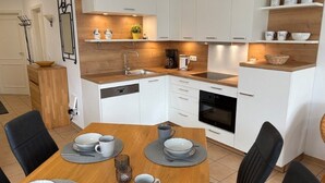 A modern kitchen with white cabinets, a wooden countertop, and a dining area with a table set for four. - (91) 3-room apartment south-facing balcony - apartment house Atlantic (Kühlungsborn)