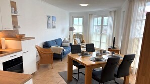 A modern living room with a sofa, dining table, and chairs. There is a kitchen area with cabinets and a countertop. - (91) 3-room apartment south-facing balcony - apartment house Atlantic (Kühlungsborn)