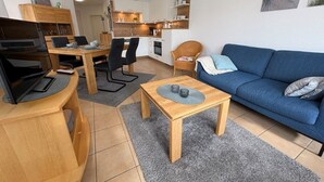 A modern living room with a blue sofa, wooden coffee table, and a dining area with a wooden table and chairs. - (91) 3-room apartment south-facing balcony - apartment house Atlantic (Kühlungsborn)
