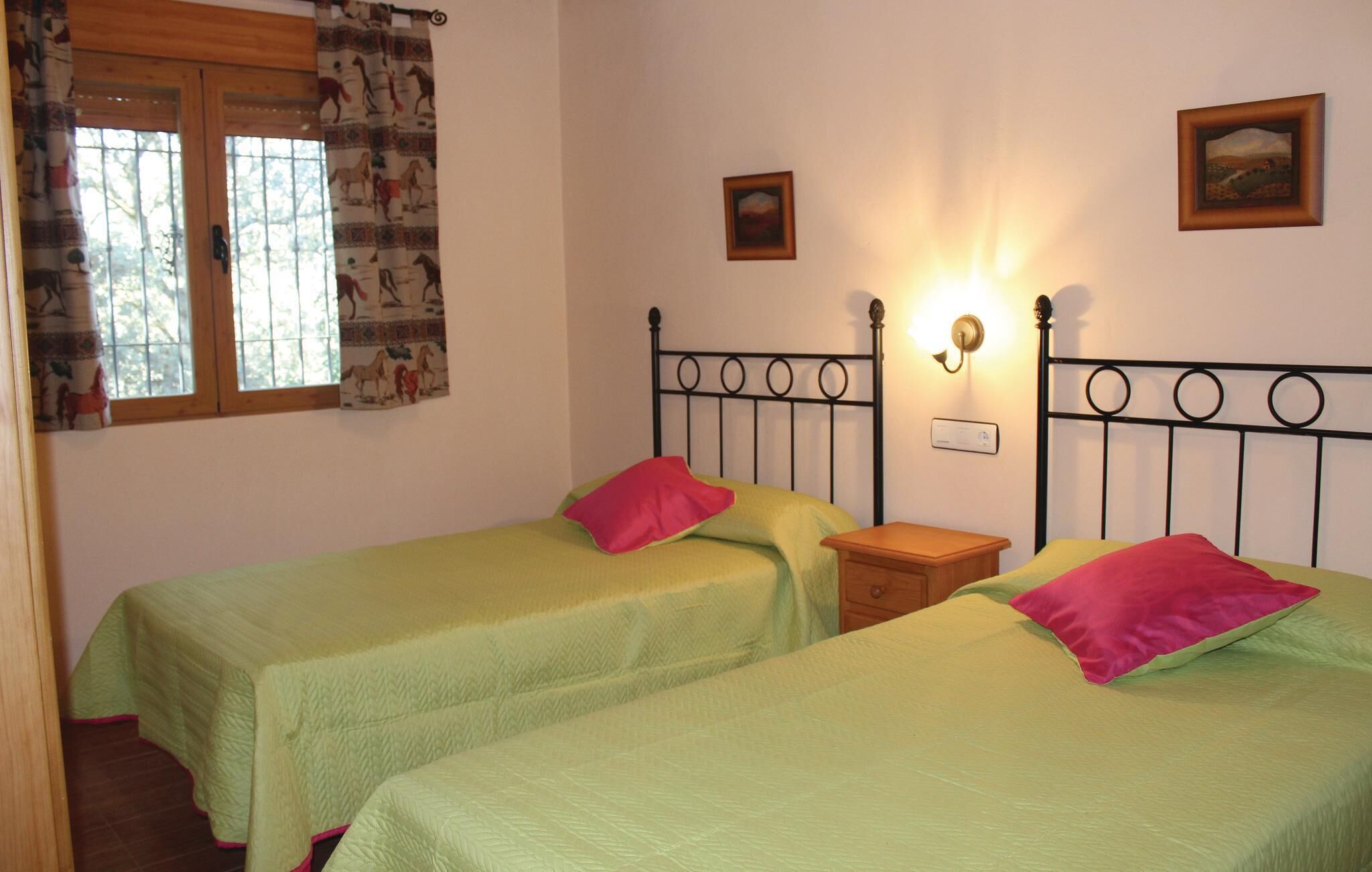 Habitació amb dos llits individuals, cadascun amb un estovat verd i un coixí rosa. Hi ha una finestra amb cortines estampades, una làmpada penjada a la paret i dos quadres emmarcats.