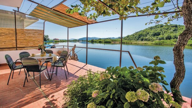 Een terras buiten met een tafel en stoelen, met uitzicht op een kalm meer en weelderige begroeiing.