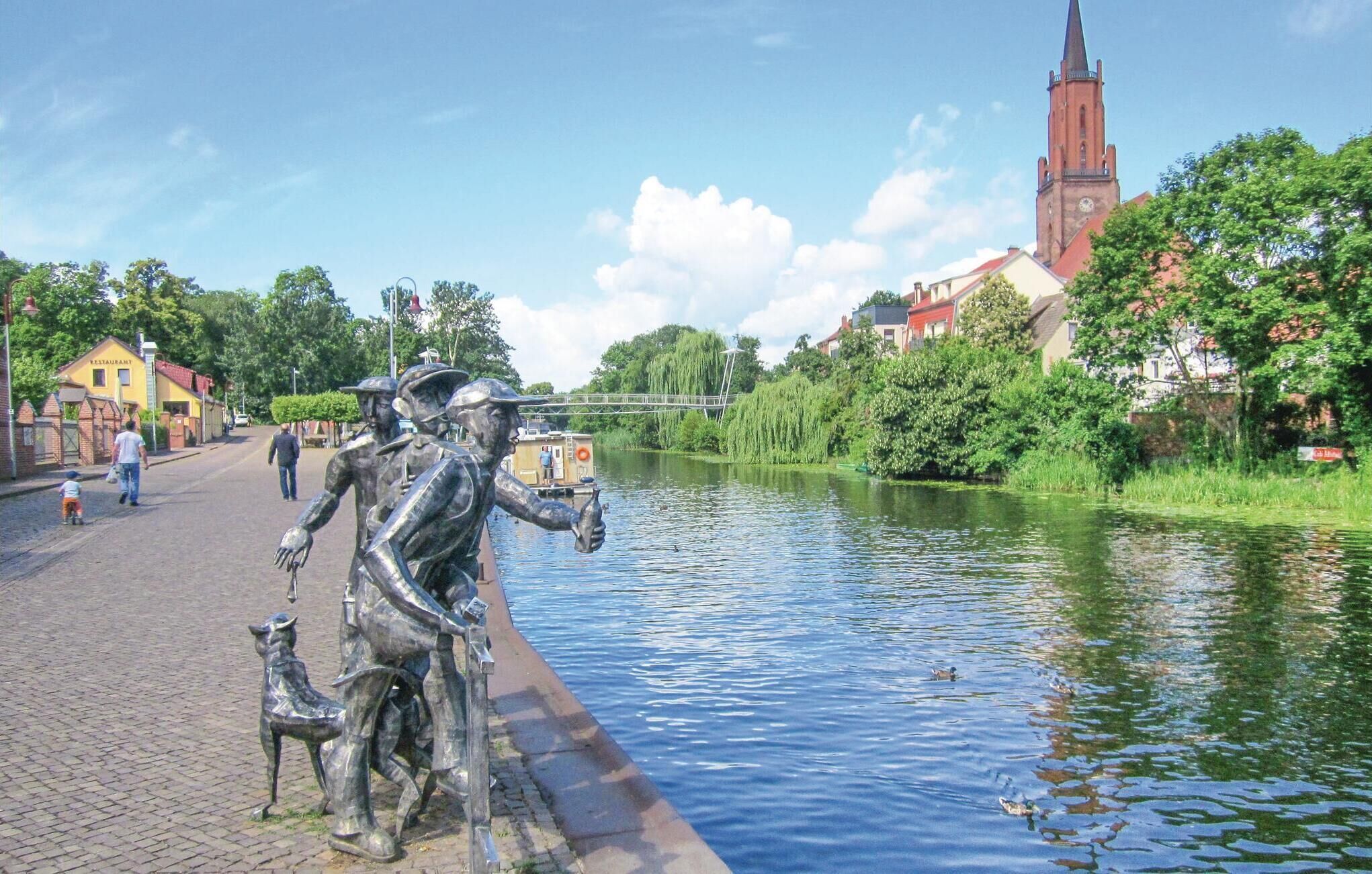 Une statue représentant un homme accompagné d’un chien, avec une rivière, un pont et des bâtiments en arrière-plan.