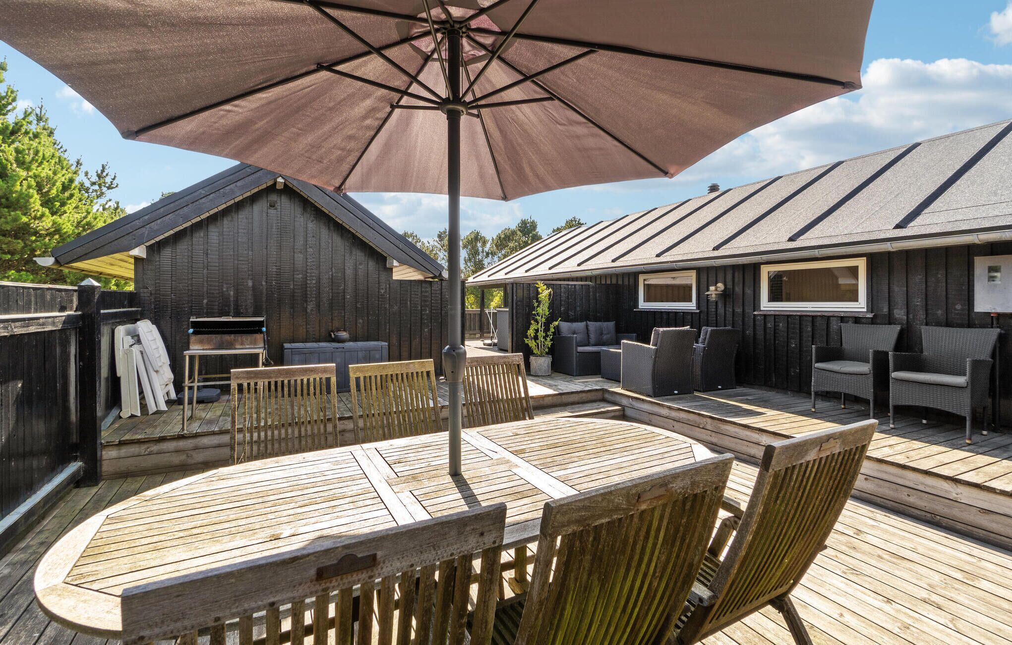 Eine Holzterrasse mit Tisch und Stühlen, einem Grill und einem großen Sonnenschirm.