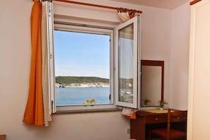 A room with a window overlooking a body of water and a beach, a wooden desk with a chair, and a mirror.