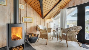 Ein gemütliches Zimmer mit Kaminofen, Korbsesseln, einem Esstisch und einem großen Fenster mit Blick auf eine verschneite Landschaft.