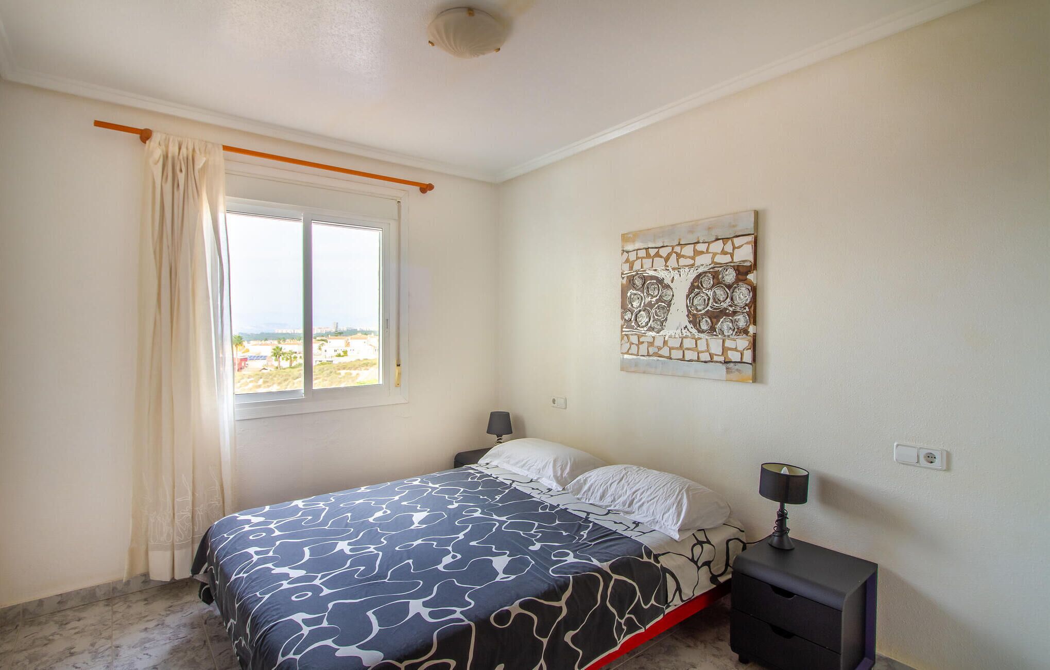 A bedroom with a bed, a nightstand, a lamp, a window with curtains, and a wall art piece.