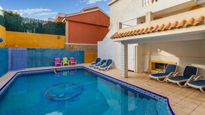 Una piscina con baldosas azules y un borde decorativo, rodeada de sillones de descanso y una pequeña zona junto a la piscina con un muro blanco y un edificio rojo al fondo.