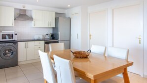 Cocina compacta con muebles blancos, un microondas, un tostador y una lavadora. Hay una mesa de madera con cuatro sillas y una cesta tejida.