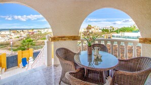 Un balcón con mobiliario de mimbre y una mesa, con vistas a un pueblo costero con edificios y vegetación.