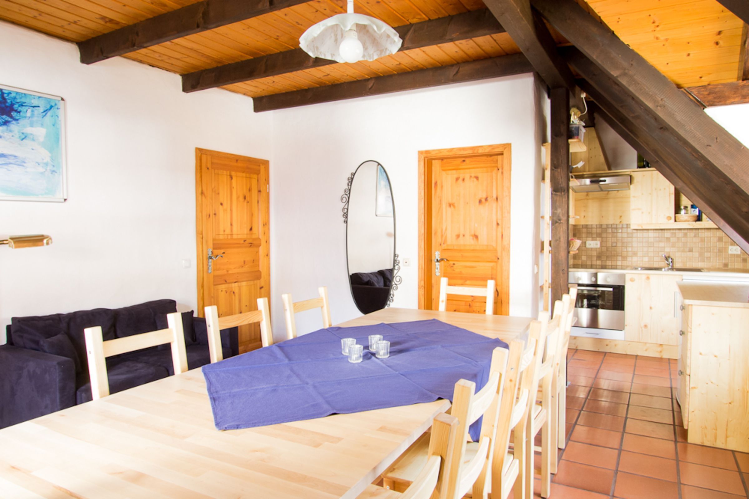 A dining area with a wooden table, chairs, and a blue tablecloth. There is a kitchen visible in the background.
