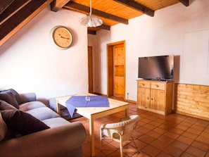 Ein gemütliches Wohnzimmer mit einer Couch, einem Tisch, einem Korbstuhl, einem Fernseher auf einer Holzkommode und einer Uhr an der Wand.