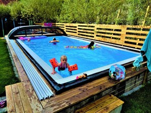 Ein Schwimmbecken mit Holzterrasse, umgeben von einem Zaun und Grünpflanzen.