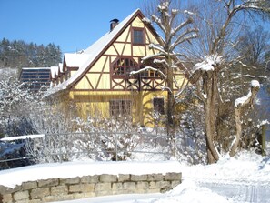 Ein traditionelles Fachwerkhäuschen mit geneigtem Dach, umgeben von schneebedeckten Bäumen und einer Steinmauer.