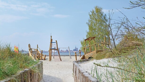 Un sentiero sulla spiaggia conduce a una struttura in legno con altalene, circondata da sabbia e vegetazione.