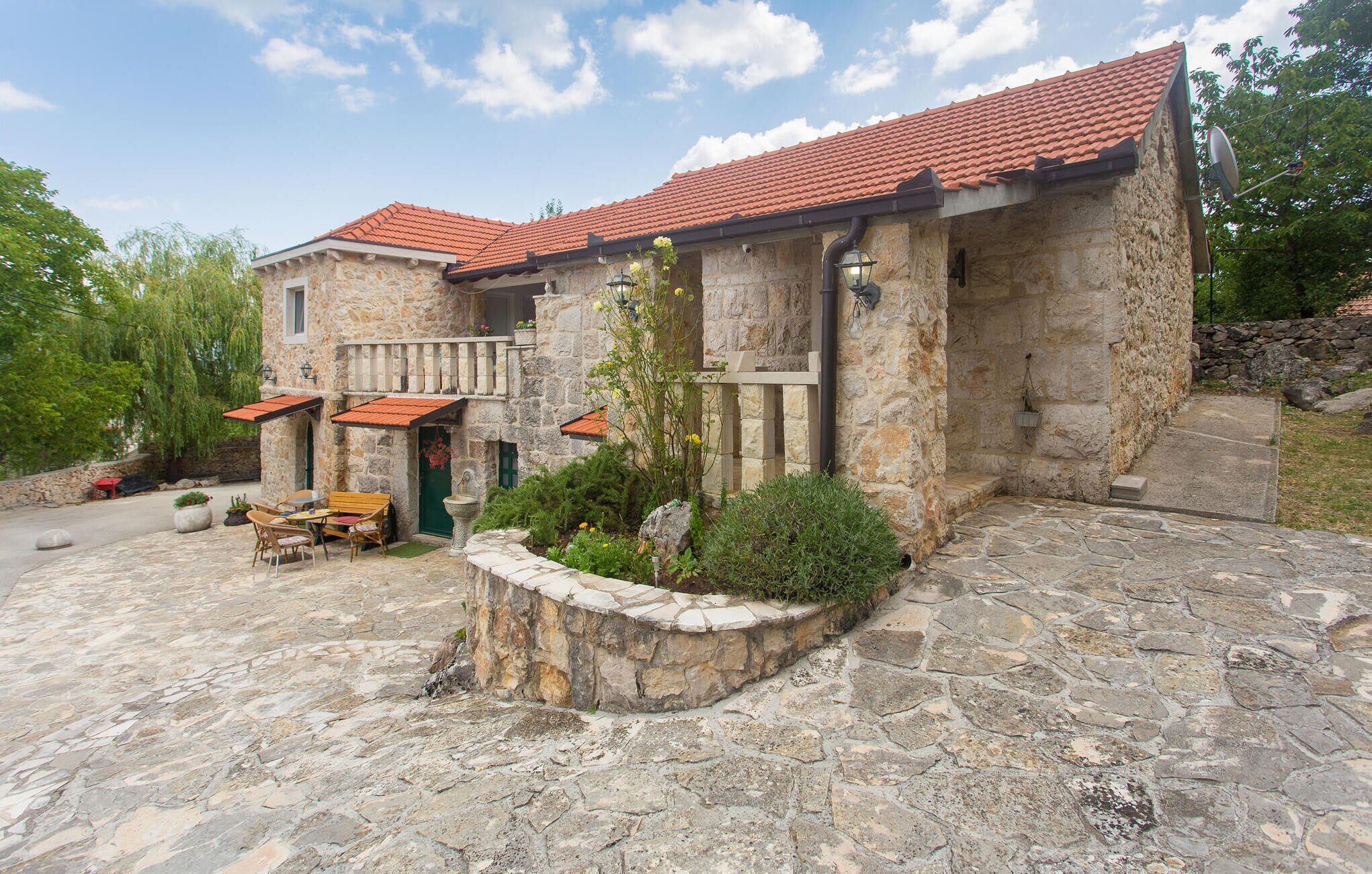 A stone building with a red-tiled roof, a small outdoor seating area with tables and chairs, and a stone pathway leading to the entrance.