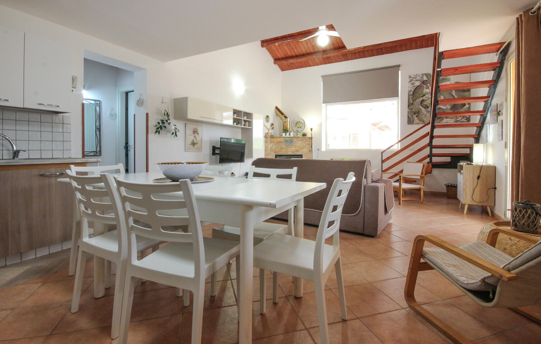 A modern kitchen with white cabinetry, a dining area with white chairs, a sofa, and a staircase with wooden railings.