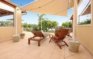 A covered outdoor seating area with two wooden lounge chairs, a stone planter, and a view of a swimming pool and greenery. - Awesome home in Menfi AG (Menfi AG)