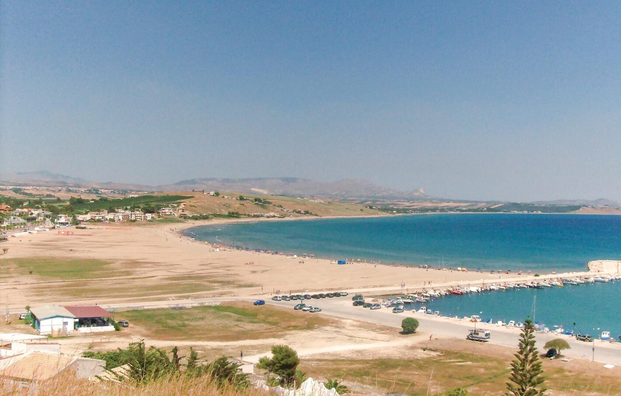 A coastal town with a beach, a marina, and a road leading to a building with a red roof.