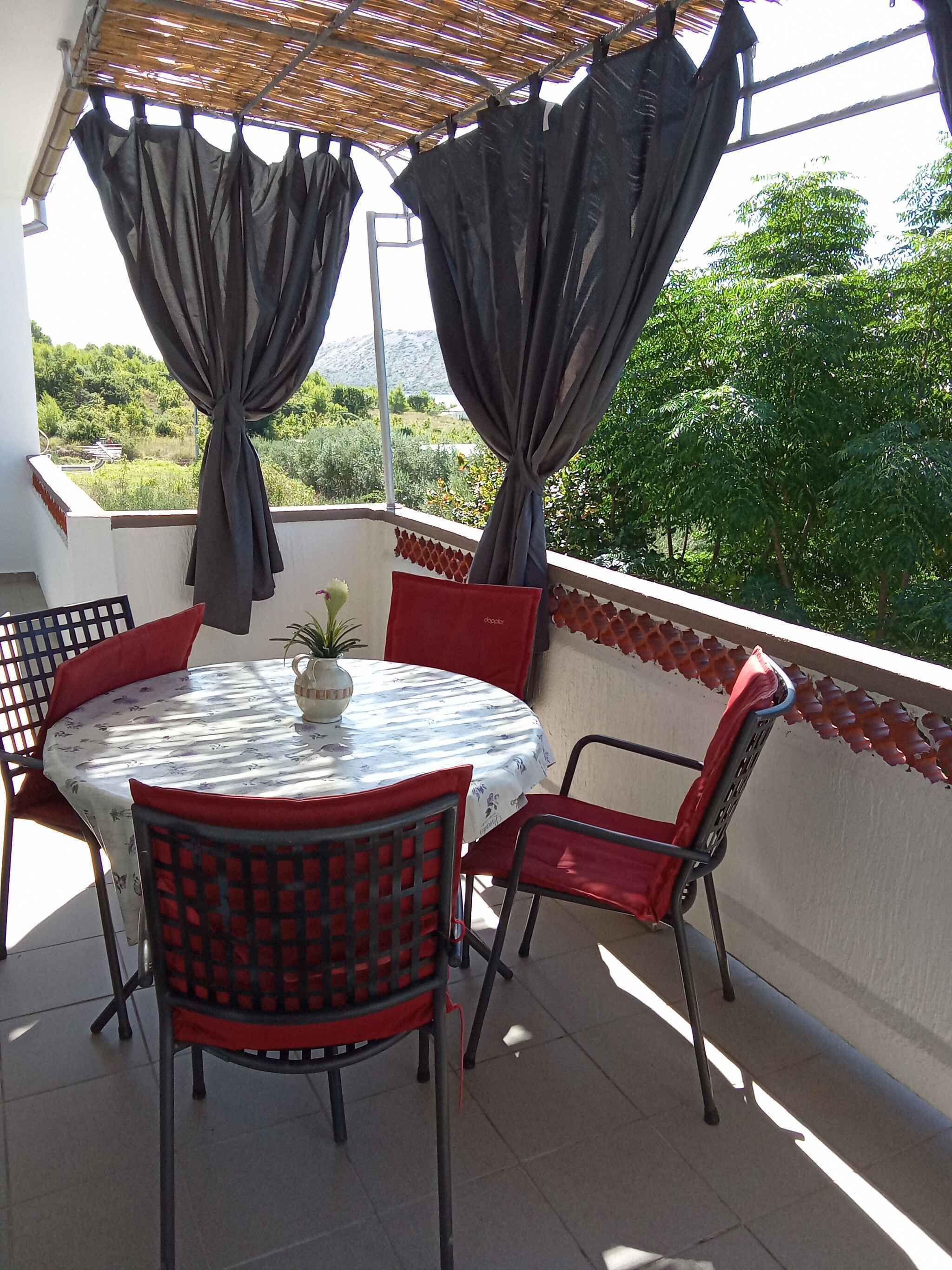 A round table with a floral tablecloth, surrounded by red chairs with black frames, on a balcony with a view of greenery and mountains.