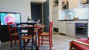 Un coin repas avec une table et des chaises en bois, un téléviseur diffusant un dessin animé, et une cuisine avec des meubles et des appareils électroménagers blancs.