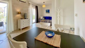 Un espace repas moderne avec une table sombre, des chaises blanches et un bol bleu. On y trouve un meuble blanc avec une lampe, un canapé avec des coussins bleus et un tableau accroché au mur.