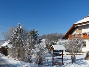 A snow-covered landscape with a wooden sign indicating a lodging area, surrounded by snow-laden trees and a building with a sloped roof.