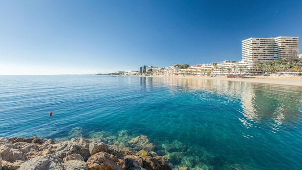 A coastal view with clear blue waters, a rocky shoreline, and a beachfront hotel with multiple floors and balconies.
