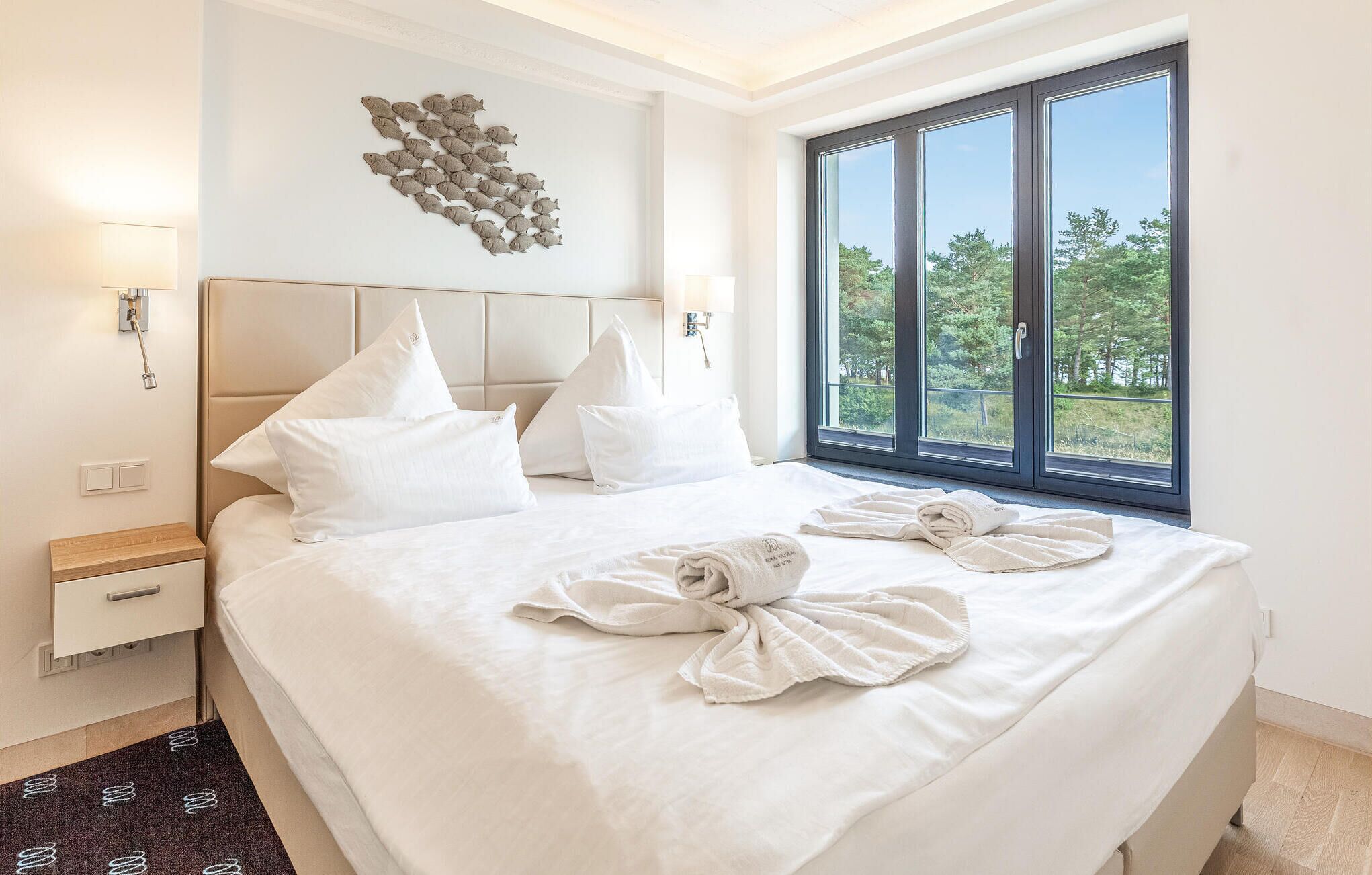Hotelkamer met groot bed, wit bedlinnen, hoofdbord, een wandlamp en een raam met uitzicht op bomen. Op het bed staat ook een handdoeksculptuur.