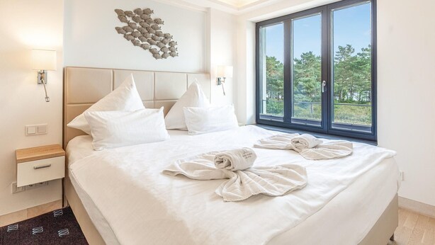 A hotel room with a large bed, white bedding, a headboard, a wall-mounted light fixture, a window with a view of trees, and a towel art on the bed.