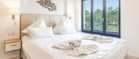 A hotel room with a large bed, white bedding, a headboard, a wall-mounted light fixture, a window with a view of trees, and a towel art on the bed.
