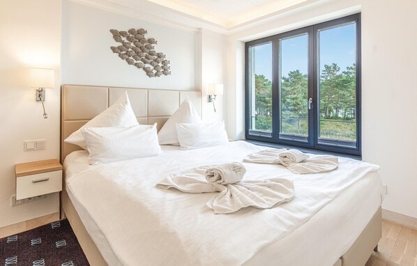 A hotel room with a large bed, white bedding, a headboard, a wall-mounted light fixture, a window with a view of trees, and a towel art on the bed.
