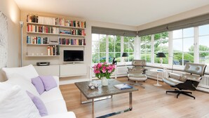 A spacious living room with a white sofa, a coffee table, a bookshelf, and a television. Large windows provide a view of greenery outside. - Hiidbloom-Hüs from A&M - Hiidbloom-Hüs (Kampen (Sylt))