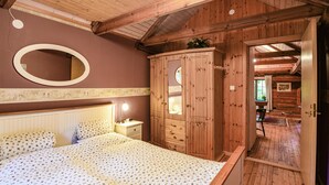 A bedroom with a wooden ceiling, a bed with floral bedding, a bedside table with a lamp, a wardrobe, and a mirror.