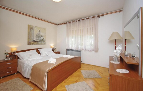 Une chambre à coucher avec un lit en bois, du linge de lit blanc, une table de chevet avec une lampe, une commode avec un miroir et une fenêtre avec des rideaux légers.